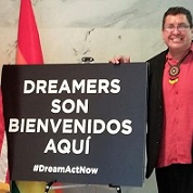 Image description: Picture of Hector outside of Senator Kamala Harris office in Washington DC. He is standing next to a sign that reads ‘DREAMERS SON BIENVENIDOS AQUI #DreamActNow” There is a pride rainbow flag next to the sign. Hector is wearing black pants and a black jacket. He is wearing a salmon color dress shirt and is wearing a Native American tribal medallion. Hector is smiling at the camera and wearing glasses. 